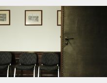 Ein Warteraum mit mehreren gepolsterten Stühlen vor einer Wand mit Holzverkleidung im unteren Bereich. An der weiß gestrichenen Wand hängen drei gerahmte Schwarz-Weiß-Bilder; rechts im Bild ist eine geöffnete Tür mit dunkler Lederoptik sichtbar.