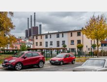 Mehrfamilienhaus in einer Wohngegend mit pastellfarbenen Fassaden, davor geparkte Autos auf der Straße. Im Hintergrund sind Industrieschornsteine zu sehen, und der Herbst ist erkennbar durch gelbes Laub an den Bäumen.