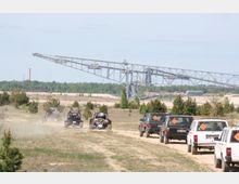 Mehrere Geländewagen und Offroad-Fahrzeuge fahren auf einer staubigen Piste in einer Landschaft mit vereinzelten Bäumen, im Hintergrund ist eine große Förderbrücke und ein bewaldeter Horizont zu sehen.