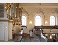 Innenraum einer historischen Kirche mit einer prunkvollen Orgel, geschmückt mit goldenen Verzierungen. Im Hintergrund sind hohe Rundbogenfenster, weiße Wände mit klassizistischem Dekor, Holzstühle sowie Emporen mit hölzernen Sitzreihen und schmiedeeisernen Geländern zu sehen.