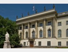 Neoklassizistische Fassade eines Universitätsgebäudes mit großen Säulen, verzierten Fenstern und Statuen auf dem Dach; im Vordergrund eine Statue vor grünen Bäumen.