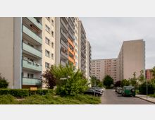 Mehrstöckige Wohnhäuser mit Balkonen und bunten Fassaden in einer städtischen Umgebung. Im Vordergrund sind eine schmale Straße, geparkte Autos und Bäume zu sehen.