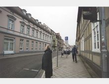 Straßenszene mit historischen Gebäuden, darunter ein rosa verputztes Haus mit weißen Fenstern und Gauben auf der linken Seite sowie ein Fachwerkgebäude rechts. Gehweg, parkende Autos und Fußgänger sind erkennbar, dazu ein Verkehrsschild und eine Ampel im Vordergrund.