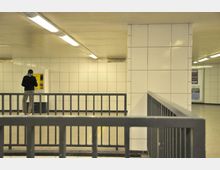 Unterirdischer, gefliester Bereich mit weißer Wandverkleidung und Metallgeländern; im Hintergrund steht eine Person vor einem gelben Automaten.