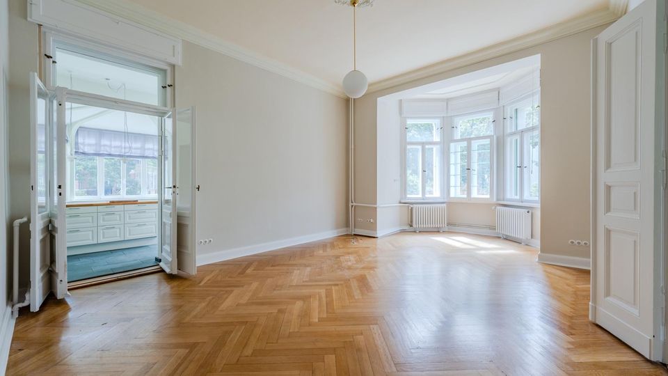 Heller, leerer Altbau-Wohnraum mit Fischgrätparkett, hohen Decken mit Stuck und weißer Pendelleuchte. Große Erkerfenster mit Heizkörpern darunter sowie Flügeltüren führen in einen angrenzenden Raum mit Einbauschränken.