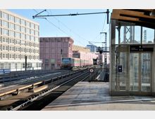 Städtischer S-Bahnhof mit Gleisanlage, einem einfahrenden Zug und einem modernen gläsernen Aufzug. Im Hintergrund sind ein rosa Gebäudekomplex mit Geschäften und eine Reihe von Oberleitungen sichtbar.