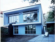 Zweistöckiges modernes Gebäude mit großer Glasfassade und rechteckigen Formen. Vor dem Gebäude liegt eine gepflasterte Terrasse, und im Hintergrund sind Bäume und Grünflächen sichtbar.