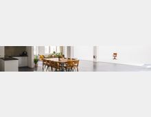 Ein großer, heller Raum mit weißen Wänden und einem dunklen Boden, ausgestattet mit einem langen Holztisch und mehreren Stühlen. Links befindet sich eine kleine Küchenzeile, während rechts ein einzelner Stuhl vor einer leeren weißen Fläche steht; im Hintergrund sind Fenster und Pflanzen sichtbar.