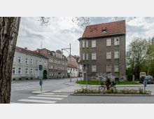 Straßenszene mit einem grauen, dreistöckigen Wohngebäude mit roten Dachziegeln im Vordergrund, umgeben von weiteren Mehrfamilienhäusern. Ein Fahrradfahrer fährt nahe einem Fußgängerüberweg, und im Hintergrund sind Bäume und Büsche zu sehen.