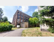 Backstein-Industriegebäude mit großen Fensterflächen, umgeben von Bäumen und Büschen, auf einem Hof mit geteerten Wegen und Grasflächen; seitlich ist ein kleineres graues Nebengebäude sowie ein geparkter Anhänger zu sehen.