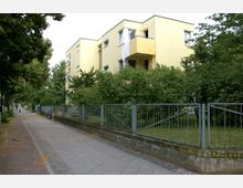 Mehrstöckiges, gelb gestrichenes Wohngebäude mit flachen Dächern und kleinen Balkonen, umgeben von Bäumen und Büschen. Im Vordergrund befindet sich ein Gehweg neben einer umzäunten Grünfläche.