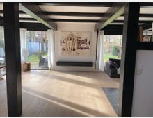 Helles Wohnzimmer mit sichtbaren Holzbalken und großen Glasfenstern, die auf einen Garten mit Rasenfläche und Terrasse blicken. Der Raum ist schlicht eingerichtet mit einem modernen Wandregal, einem großformatigen Wandbild und einem Holzboden.