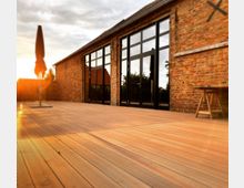 Ziegelstein-Gebäude mit großen, schwarzen Glastüren und Fenstern, davor eine großzügige Holzterrasse bei Sonnenuntergang. Links ein geschlossener Sonnenschirm, im Hintergrund Bäume und ein Zaun.