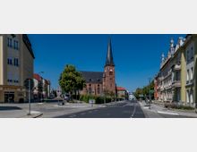 Straßenszene in einer Kleinstadt mit einer roten Backsteinkirche und spitzem Kirchturm auf der linken Straßenseite, flankiert von Bäumen. Rechts stehen mehrstöckige Wohnhäuser im historischen Stil, und im Vordergrund sind eine Apotheke und Ampeln sichtbar.