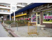 Kleines Einkaufszentrum mit Backsteinfassade und blauer Überdachung, das verschiedene Ladenfronten wie ein Café („Am Brückenbogen“) und ein Friseursalon („Frisch“) zeigt. Davor befinden sich ein Fahrradständer, bepflanzte Begrenzungen und ein gepflasterter Gehweg.