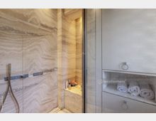 Moderne Dusche aus Natursteinoptik mit horizontalen Streifen; ausgestattet mit Wandarmaturen und einer Handbrause. Rechts befinden sich eingebaute Regalfächer mit aufgerollten Handtüchern.