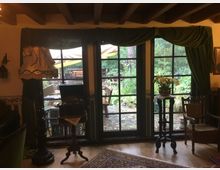 Wohnraum mit rustikalem und vintage-inspiriertem Dekor, einschließlich eines Kronleuchters, dunkelgrüner Vorhänge und einer Lampe mit verspieltem Stoffschirm. Große Fenster mit Blick auf eine Terrasse und einen grünen Garten im Hintergrund.