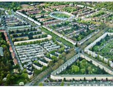 Luftaufnahme einer Wohnsiedlung mit vielen mehrstöckigen Wohngebäuden, angeordnet in Blockform und umgeben von grünen Bäumen, Gärten und Parkflächen. Im Hintergrund erstreckt sich ein großer, dicht bewachsener Wald.