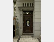 Eingangstür eines historischen Gebäudes mit verzierten Holzelementen und Glaseinsätzen, umrahmt von hellen Steinwänden. Ein gepflasterter Weg führt zur Tür, die von mehreren kleinen Fenstern oberhalb abgeschlossen wird.