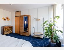 Schlafzimmer mit blauer Teppichboden, einem Holzbett (nicht vollständig sichtbar), einer Kommode aus Holz und Metall, einem Holzkleiderschrank mit Spiegel, einem schwarzen Kleiderständer, einem großen Zimmerpflanze in einem Topf sowie weißen Vorhängen an bodentiefen Fenstern.