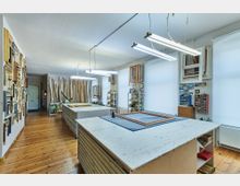 Ein geräumiger Werkstattraum mit Holzboden, großen Arbeitstischen in der Mitte und langen Leuchtstofflampen an der Decke. Wände sind mit gerahmten Bildern und Holzleisten dekoriert, während Regale mit Werkzeugen und Materialien an den Seiten stehen.