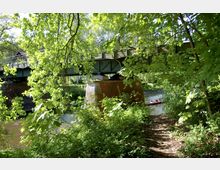 Eine grüne Flusslandschaft mit einer Stahlbrücke, die von massiven Backsteinpfeilern gestützt wird. Im Vordergrund sind dichte Bäume und Büsche sichtbar, und ein rotes Kanu mit zwei Personen befindet sich auf dem Fluss unter der Brücke.