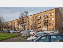 Wohnblock im Stil der Nachkriegsarchitektur mit braun verputzten Fassaden, mehreren Balkonen und weißen Fenstern. Im Vordergrund eine Reihe geparkter Autos, eine grüne Rasenfläche und ein Straßenschild mit der Aufschrift "Trientje Straße".