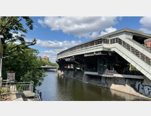 Eine erhöhte Bahnhaltestelle mit einem modernen, stählernen Dach, gelegen an einem Flussufer. Rechts sind Treppen mit verglasten Geländern und Graffitis an den Betonwänden sichtbar, während im Hintergrund eine Brücke und Gebäude erkennbar sind.