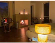 Gemütliches Wohnzimmer mit Holzboden, zentralem Kaminfeuer und Kaminsims mit Kerzen und kleinen Skulpturen. Große Fensterfronten, ein niedriges Sofa sowie ein Beistelltisch und Stühle ergänzen die warm beleuchtete Einrichtung.