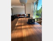 Heller Wohn- und Essbereich mit großem Holztisch im Vordergrund, schwarzem Kerzenhalter-Set und Pendelleuchten an der Decke. Im Hintergrund stehen Sofa und Couchtisch neben einem modernen Kamin; rechts führen bodentiefe Fenster mit Vorhängen zu einem grünen Garten bzw. Balkon.