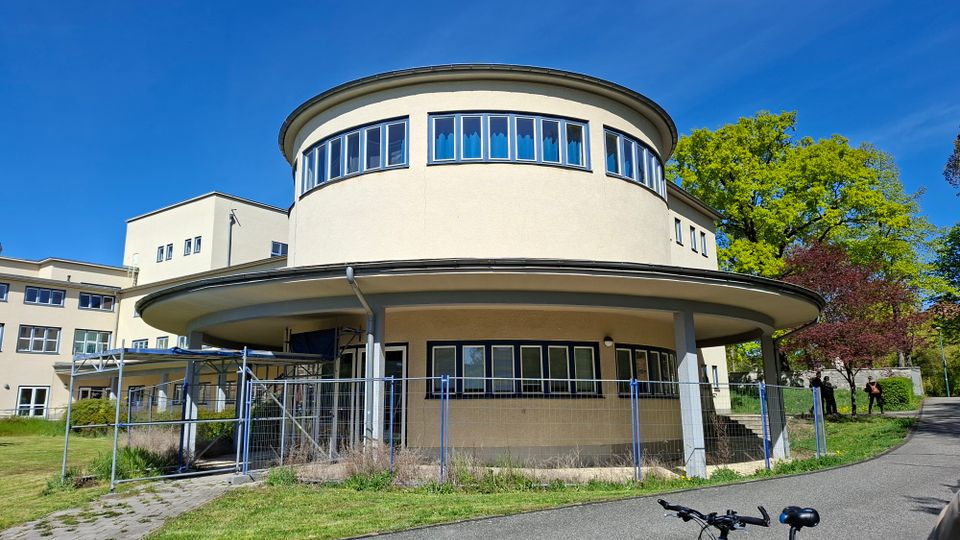 Rundes, zweigeschossiges Gebäude mit flachem, weit auskragendem Dach und umlaufendem Fensterband im Obergeschoss, vor einer größeren Gebäudefront. Im Vordergrund stehen ein Bauzaun und ein überdachter Eingangsbereich; daneben verläuft ein asphaltierter Weg mit Rasenflächen und Bäumen.