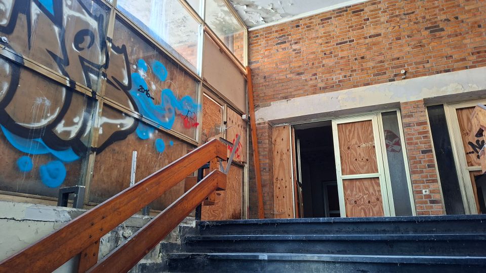 Verlassener Eingangsbereich eines Gebäudes mit Backsteinwänden, beschädigter Decke und einem Treppenaufgang mit Holzgeländer. Große Fensterflächen und Türen sind mit Sperrholz vernagelt und mit Graffiti besprüht.