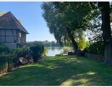 Grüne Gartenlandschaft mit Rasenfläche, rechts und links von Bäumen und Büschen umgeben, im Hintergrund ein Fachwerkhaus mit rotem Ziegeldach und ein See, dessen Ufer von Bäumen gesäumt ist. Im Vordergrund ein Zaun mit einem Gartenstuhl und am rechten Uferbereich ein Steg.