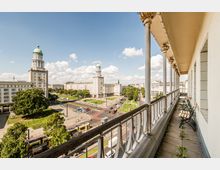 Blick von einem langen Balkon mit Säulen und Metallgeländer über eine breite Stadtstraße mit Straßenbahn, Kreuzung und Grünflächen. Gegenüber stehen zwei markante, mehrstöckige Gebäude mit runden Türmen und grünen Kuppeln vor blauem Himmel.