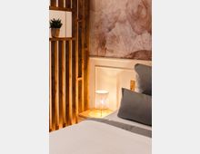 Ein gemütliches Schlafzimmer mit einem Holzgitter als Raumteiler, einer Nachttischlampe auf einem Holztisch und einer Wand mit floralem Muster. Im Vordergrund ist ein Bett mit weißer und grauer Bettwäsche zu sehen.