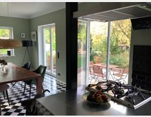 Helle Küche mit schwarz-weiß kariertem Boden, einem Holztisch mit Stühlen und einer Küchenzeile mit Gasherd im Vordergrund. Große Glas-Schiebetüren führen auf eine Terrasse mit Holzmöbeln und einem Garten im Hintergrund.