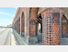 Backsteinarkaden eines historischen Gebäudes, bestehend aus roten Klinkersteinen mit dekorativen Säulen und Zierelementen. Links verläuft eine Straße mit Gehweg, im Hintergrund ist weitere Backsteinarchitektur sichtbar.