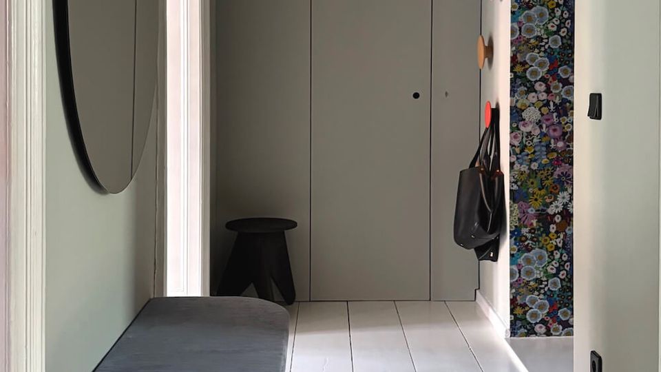 Narrow modern hallway with pale walls and white plank flooring, featuring a cushioned bench and round wall mirror on the left and a black pendant ceiling light overhead. At the far end is a flush door, with coat hooks and a colorful floral wall panel on the right.