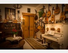 Ein gemütlicher Raum mit Vintage-Dekor, an dessen Wänden zahlreiche Gitarren und andere Saiteninstrumente hängen. Ein weißes Klavier mit offenen Notenblättern steht rechts, während links ein dunkler Holzschrank und eine Sitzgruppe mit Ledersofas und einem kleinen Tisch arrangiert sind; darüber ein Kronleuchter und Holztüren im Hintergrund.