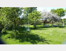 Grüne Gartenlandschaft mit blühenden Obstbäumen, einer Wiese und mehreren Bänken und Picknicktischen. Im Vordergrund steht eine dekorative, grüne Metall-Schaukel.