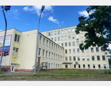 Großes, beigefarbenes Bürogebäude mit mehreren Etagen und rechteckigen Fenstern; das Gebäude hat einen L-förmigen Grundriss. Im Vordergrund befinden sich eine Grünfläche, ein Gehweg, Verkehrsampeln und ein großer Baum.