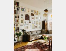 Wohnzimmer mit einem grauen Ecksofa, einer Glas-Couchtischplatte, einem roten Teppich und einer großflächigen Wandgalerie mit verschiedenen Kunstwerken. Links steht ein großes Bücherregal, und rechts sorgen große Fenster und dekorative Holzjalousien für Tageslicht.