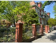 Backsteingebäude mit weißen Fensterrahmen im Stil eines historischen Mehrfamilienhauses, umgeben von Bäumen und einem dekorativen Metallzaun mit Backsteinpfeilern. Im Vordergrund verläuft ein gepflasterter Gehweg.