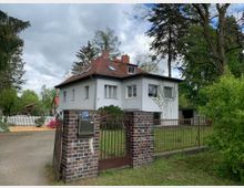 Zweistöckiges Haus mit weißer Fassade und rotem Satteldach, umgeben von einem Garten mit Rasen und Bäumen. Im Vordergrund steht ein schmiedeeisernes Tor mit gemauerten Pfeilern, das den Zugang zum Grundstück markiert.