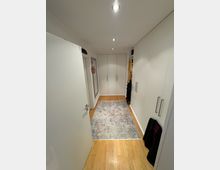 Langer, schmaler Ankleideraum mit weißen Einbauschränken an beiden Seiten, hellbraunem Holzfußboden und einem gemusterten Teppichläufer in der Mitte. Die Decke ist mit Einbauspots beleuchtet, und im Hintergrund befindet sich eine Tür oder ein weiterer Schrankabschluss.