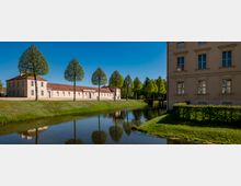 Canal bordered by grassy banks and a row of evenly spaced, trimmed trees, with long beige buildings topped by red tile roofs along the water. A larger beige building with tall windows frames the right side, and a small bridge crosses the canal in the distance under a clear blue sky.