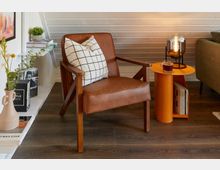 Wohnzimmer mit einem braunen Ledersessel, der mit einem schwarz-weiß karierten Kissen dekoriert ist. Daneben steht ein orangefarbener Beistelltisch mit einer Glaslampe und einer Tasse Tee, im Hintergrund ein weißes Regal mit Büchern und Pflanzen.