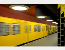 Eine Aufnahme einer U-Bahn-Station mit gelben Kacheln und moderner Gestaltung. Ein gelber U-Bahn-Zug fährt an der Bahnsteigkante vorbei, die Wand im Hintergrund ist rot mit grafischen Mustern, und runde Deckenleuchten hängen über dem Bahnsteig.