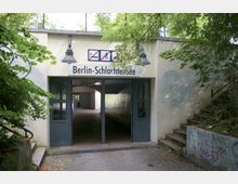 Eingang zum S-Bahnhof Berlin-Schlachtensee unter einer kleinen Unterführung, umgeben von Bäumen und Vegetation. Der Eingang zeigt ein Schild mit dem Stationsnamen, flankiert von grauen Türen und begleitet von steinernen Treppen auf der rechten Seite.