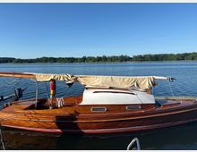 Holzsegelboot mit zusammengebundenem, beige abgedecktem Mast liegt an einem Steg auf einem ruhigen See. Im Hintergrund sind eine bewaldete Uferlinie und ein klarer blauer Himmel zu sehen.